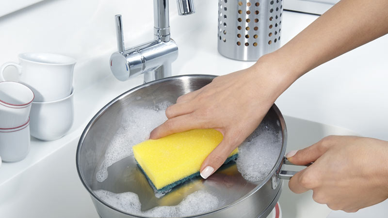 Grande plano de mão a lavar uma caçarola de inox no lava-loiça, usando um esfregão